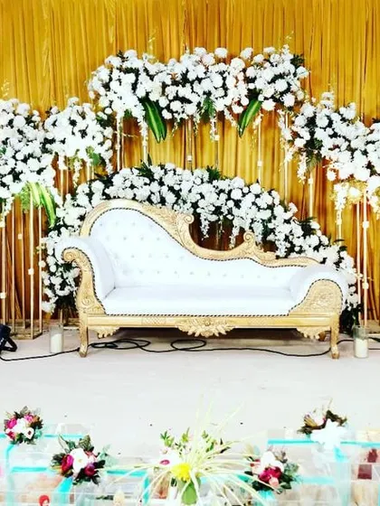 An elegant and minimalist reception stage design featuring a white chaise lounge against a backdrop of golden drapes. The stage is framed by an explosion of white flowers on tall, modern stands, creating a sophisticated and airy feel.