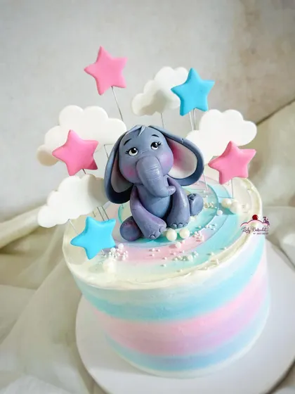 A dreamy baby shower cake with a pastel watercolor finish and a cute elephant topper sitting among the clouds and stars.