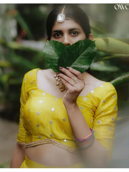 A playful and creative Haldi portrait, with the bride peeking over heart-shaped leaves.