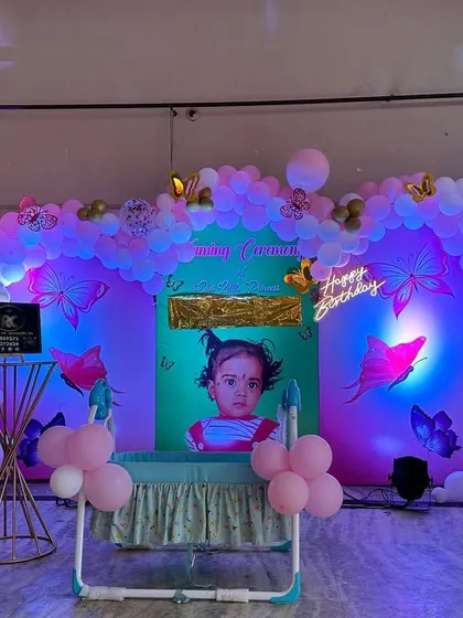This butterfly naming ceremony decoration features a custom backdrop with the baby's photo. The organic balloon arch and butterfly details are illuminated with colored spotlights to create a magical, glowing effect in the evening.