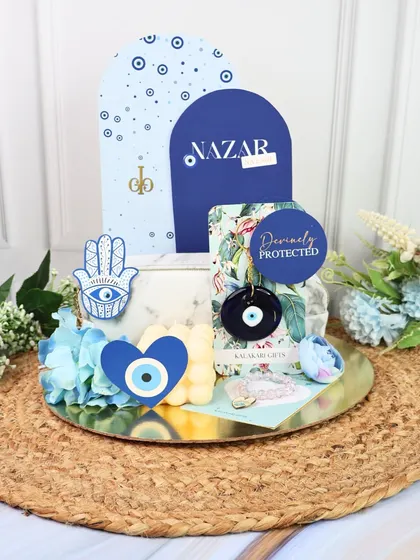 Protect your bond with this Evil Eye themed Rakhi hamper. It includes a beautiful rakhi, a bubble candle, a Hamsa hand charm, and an evil eye keychain, all arranged on a decorative gold tray.