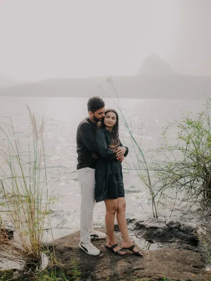 A wide shot of the couple embracing on the rocks by the lake. The misty mountains in the background add a sense of grandeur and drama to this romantic scene.