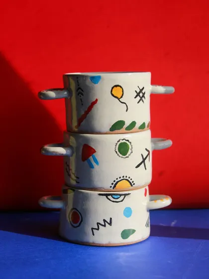 A stack of small, two handled bowls from the 'Playful Mornings' collection, decorated with colorful, abstract symbols against a bright red background.