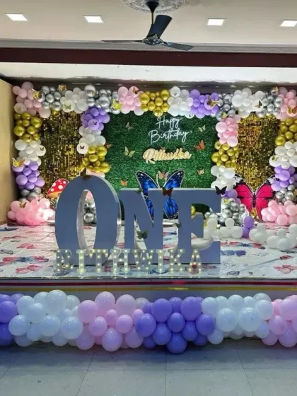 A large-scale first birthday celebration with a butterfly theme. The centerpiece is a giant "ONE" prop, set against a full-wall backdrop of greenery, shimmer panels, and pastel balloons.