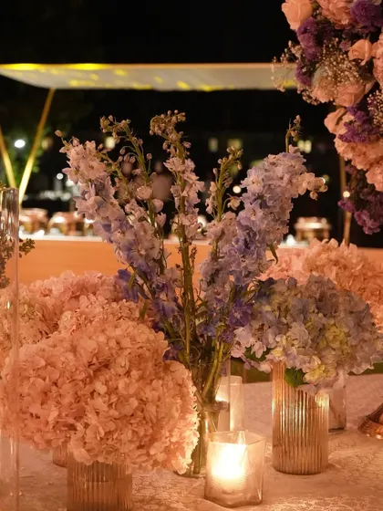 A close-up on the delicate textures of the pastel floral arrangements. Sprays of lavender delphinium and soft pink hydrangeas create a gentle, romantic look, beautifully illuminated by candlelight.