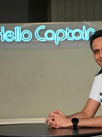 A friendly portrait of an IndiGo captain in front of a "Hello Captain" neon sign. This uses the office decor to create a unique and memorable branding shot.