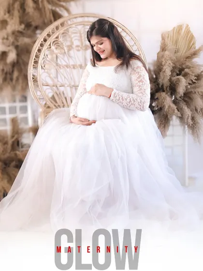 This portrait, titled 'Glow Maternity', perfectly captures the radiance of the mother-to-be. Seated in a peacock chair, she is dressed in a delicate white lace and tulle gown, looking down at her bump.