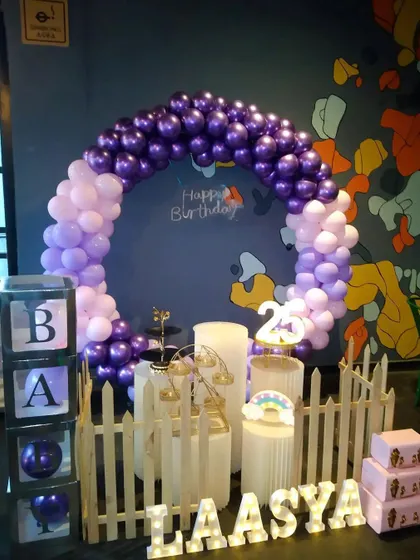 A stylish and compact birthday setup with a purple and lilac circular balloon arch. The decor is personalized with light-up marquee letters for the name "LAASYA" and includes a white picket fence and "BABY" blocks.