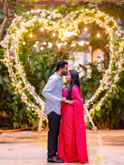 A romantic forehead kiss in front of a heart-shaped light installation. The bride's red saree adds a beautiful pop of color to this magical night shot.