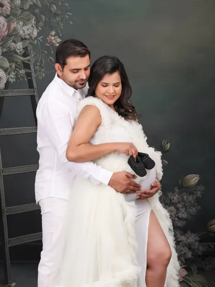 A beautiful studio portrait with a floral backdrop. The couple, dressed in white, holds a pair of baby shoes, a classic pose in pregnancy photography that signifies their growing family.