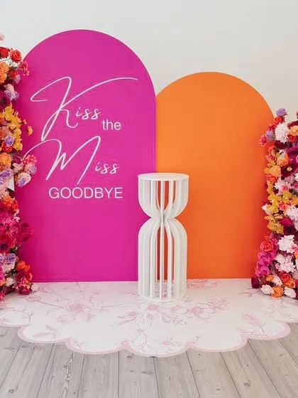 "Kiss the Miss Goodbye" with a burst of color. This vibrant setup uses a hot pink and orange backdrop with stunning floral arrangements, creating a fun and energetic photo spot.
