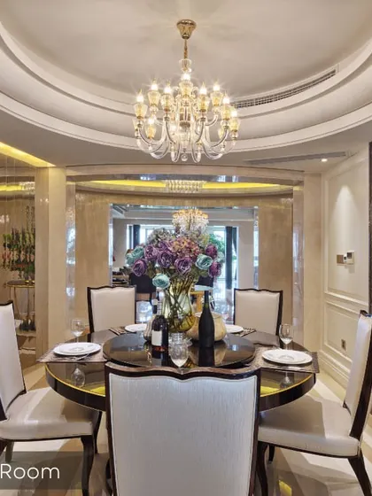 A luxurious dining room setup featuring a grand crystal chandelier and a circular false ceiling design. The space is designed for formal entertaining, with elegant chairs and a statement crockery unit against a marble-paneled wall.