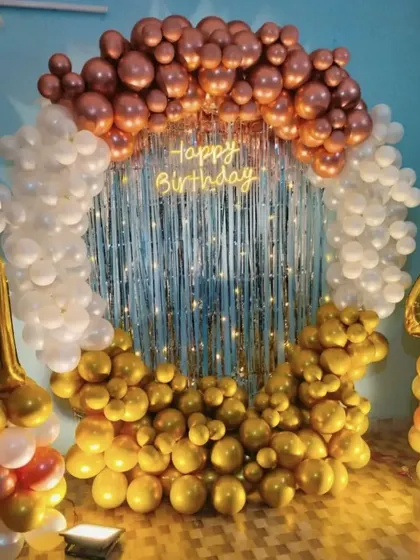 A festive birthday decoration with a circular balloon arch in rose gold, white, and gold, set against a silver fringe curtain.