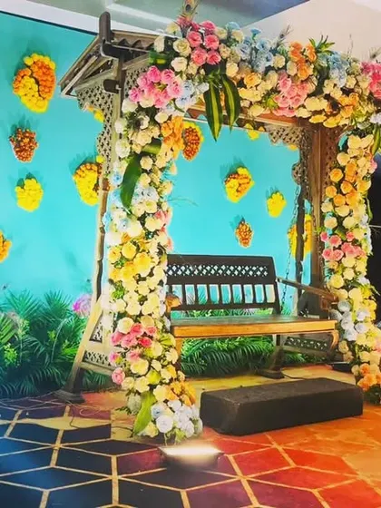 A whimsical and colorful swing setup for a Mehendi or Sangeet. The pastel floral arch and butterfly motifs on the turquoise backdrop create a fun, fairytale-like atmosphere.