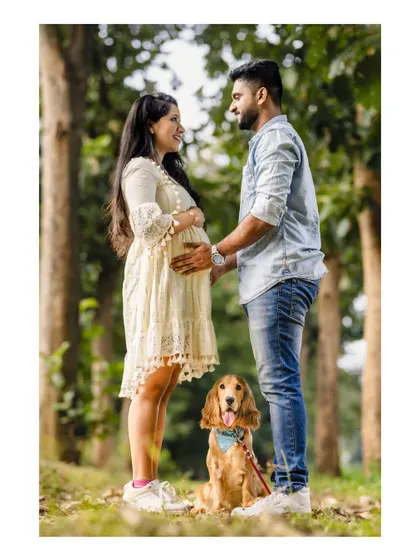 An expecting couple shares a loving gaze in a sunlit park, with their adorable dog sitting patiently at their feet. A perfect portrait of a growing family.