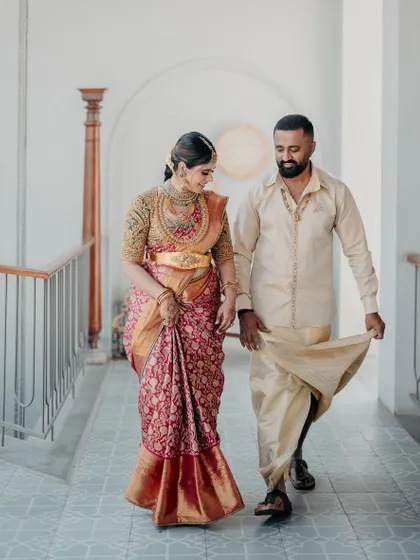 A couple walking through a corridor, hand in hand. This shot captures the beautiful drape of her Kanchipuram silk saree and the classic simplicity of his coordinated dhoti-kurta set.