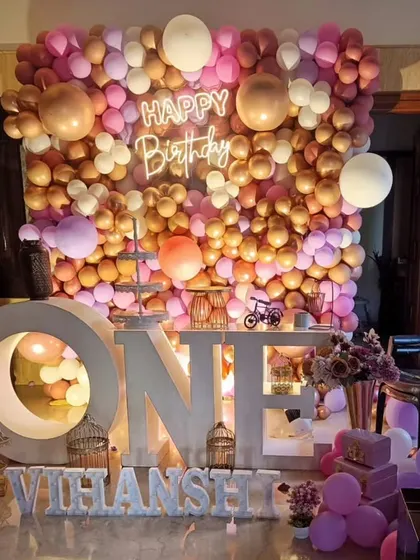 A full balloon wall in shades of pink and rose gold serves as a stunning backdrop, complemented by large "ONE" letters and a personalized name sign.