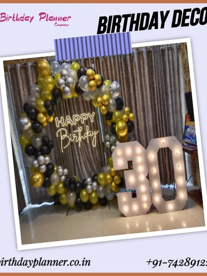 A chic setup for a 30th birthday party. The decor features a circular balloon arch in black, silver, and gold, a neon "Happy Birthday" sign, and large marquee numbers, creating a sophisticated party vibe.