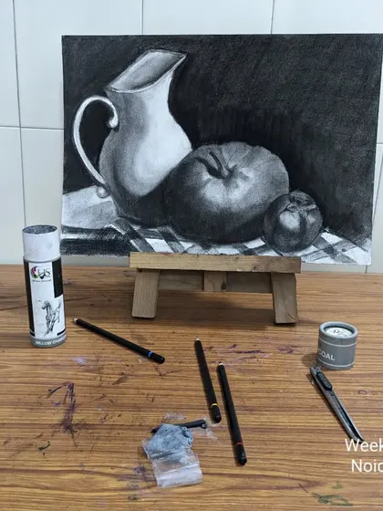 The setup for our charcoal still life class. You can see the final drawing on an easel with the charcoal sticks and blending tools used to create it, showing the tools of the trade.