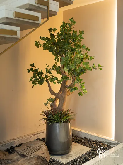 A full view of the under-staircase garden, showing the interplay of the faux tree, rocks, pebbles, and vertical accent lighting. The result is a complete and polished look.