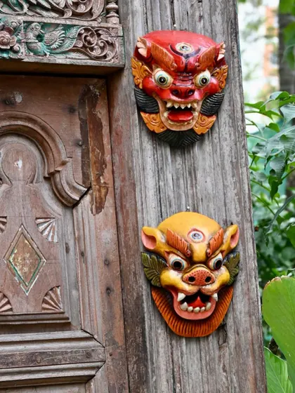 A pair of traditional Nepalese masks mounted on a rustic wooden surface. The contrast in colors and expressions showcases the variety within this folk art form, where each mask has its own story and purpose.