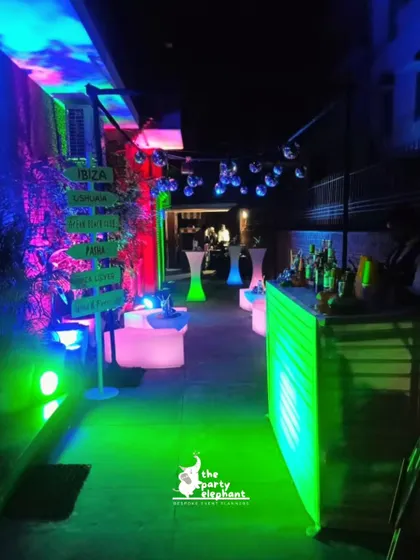 A full view of the neon-lit bar and lounge area, showing the glowing bar counter, LED tables, and custom directional sign pointing to famous Ibiza clubs.
