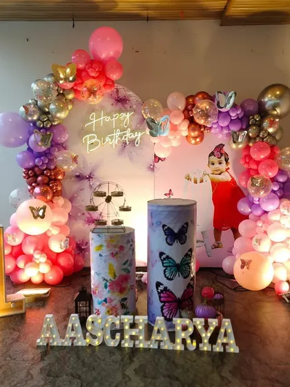 A butterfly themed first birthday decoration with a "Happy Birthday" neon sign. The setup features a rich color palette of purple and pink balloons, a light-up number '1', and personalized light-up letters for the name.