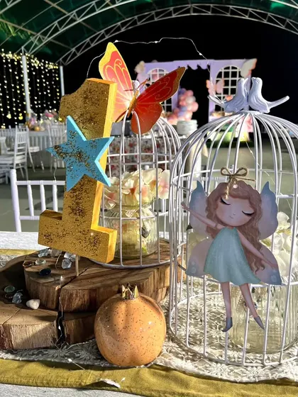 A beautiful centerpiece for the butterfly party, featuring a golden number "1," a fairy cutout in a white birdcage, and other whimsical elements on a wooden base.