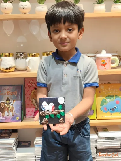 A young boy proudly displays the clay art piece he made, featuring two birds on a branch under the moon.