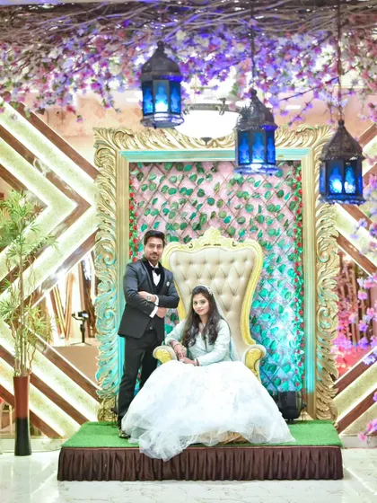 The couple poses on their grand reception stage, featuring a throne-like chair and an elaborate floral and light-filled backdrop.