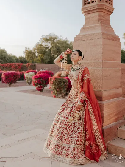 A full-length bridal portrait of Amala in her stunning red lehenga, posing against the beautiful sandstone architecture of Umaid Bhawan Palace.