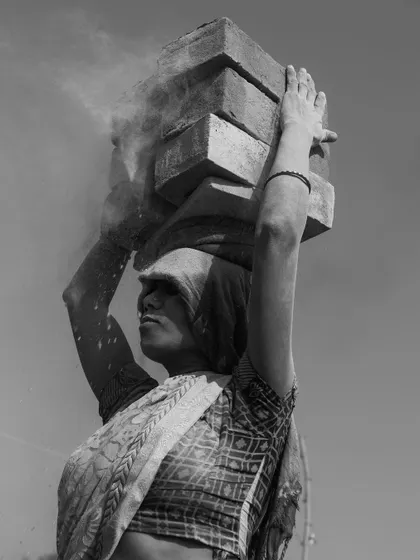 A black and white portrait of a woman carrying bricks, with dust and powder falling from her load. The image is a powerful statement on the physicality of the work.