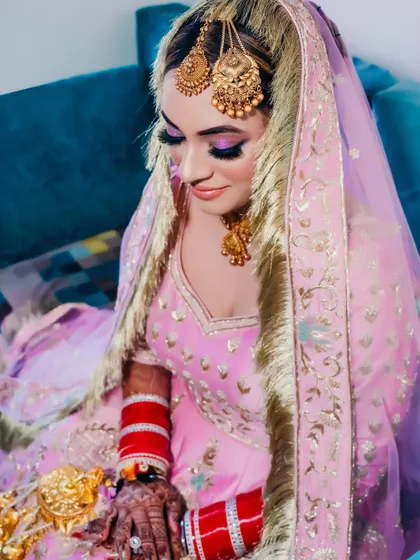 A close-up portrait of a Sikh bride in a soft pink and purple outfit. The focus is on her beautiful makeup and traditional golden jewelry.