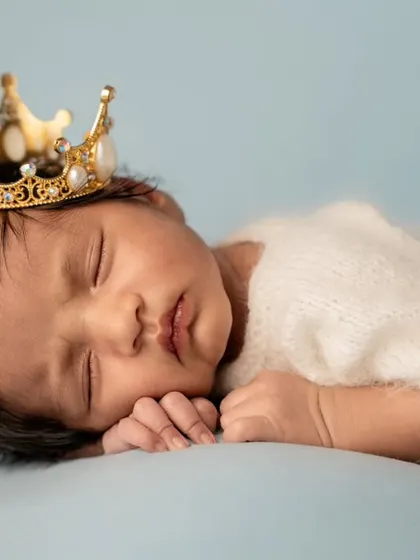 A close-up portrait of a sleeping newborn wearing a detailed gold and pearl crown, showcasing their peaceful expression.