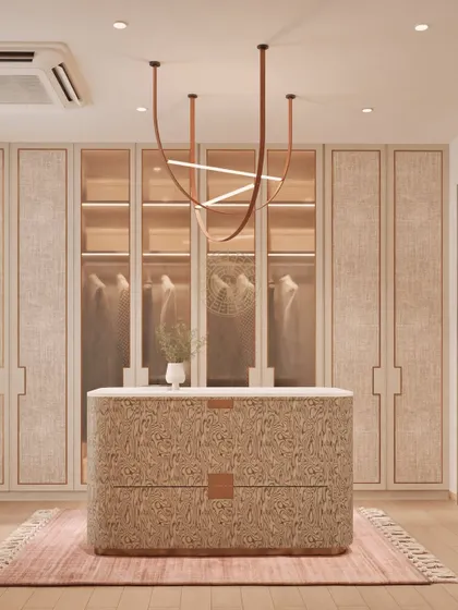 A render of a bright and airy walk-in closet with a central island made from a unique, textured material. The glass-fronted wardrobes and modern chandelier create an elegant and functional space.