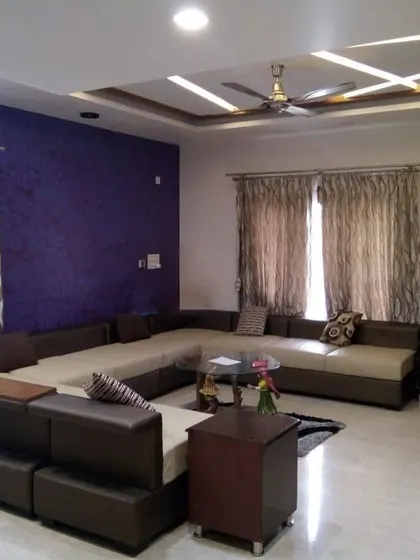 This living room makes a bold statement with a deep purple textured accent wall. A large, comfortable sectional sofa provides ample seating, while the false ceiling with strip lighting adds a modern touch to the overall design.