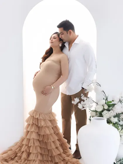 A serene and graceful portrait. The expecting mother is framed by a minimalist arch and soft florals, creating a clean, modern, and elegant look in a beautiful ruffled gown.