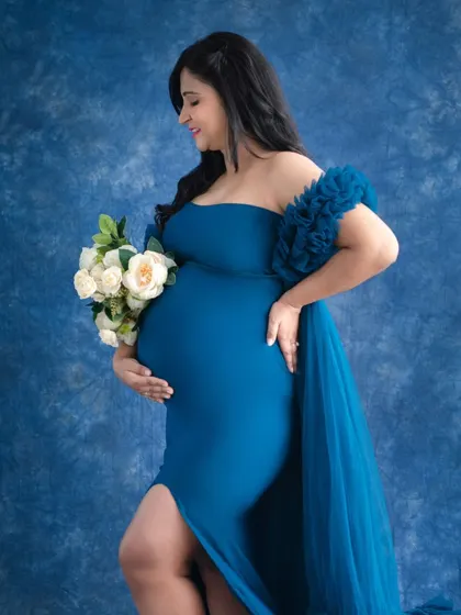 A classic profile shot that beautifully showcases the baby bump. The mother-to-be holds a bouquet, her posture elegant against a rich blue studio backdrop.