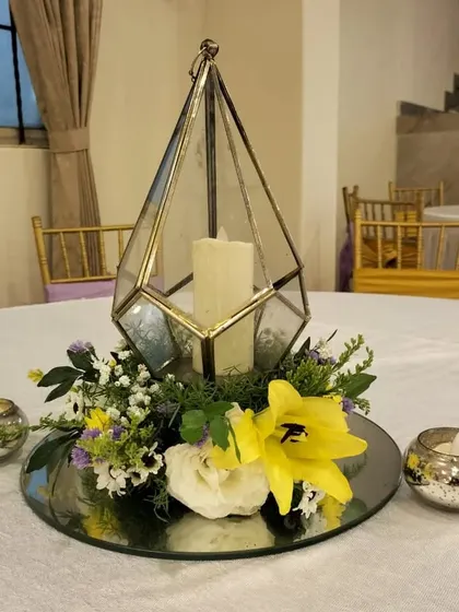 This geometric terrarium centerpiece, filled with a candle and surrounded by yellow and white flowers, was designed for an elegant anniversary dinner. The mirrored base reflects the candlelight, adding to the ambiance.