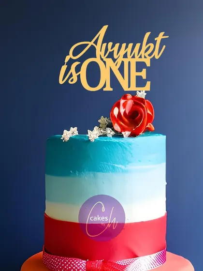 A mockup of the "Avyukt is ONE" topper on a colorful tiered cake, demonstrating how a simple gold topper can complement a vibrant cake design.