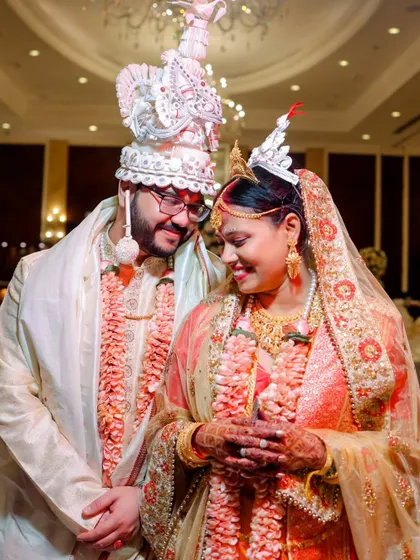 A sweet, close-up portrait of a Bengali couple on their wedding day. Their smiles and traditional headwear (topor and mukut) are the focus.
