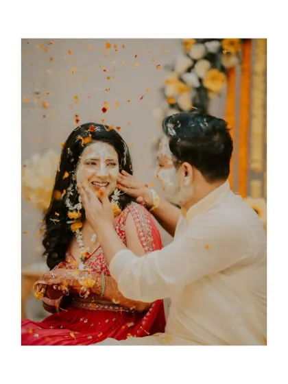 A candid shot of a couple during their haldi, with the groom applying paste to the bride's face as petals fly.