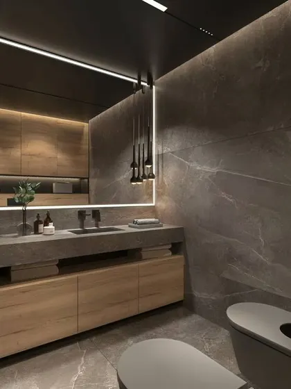 A sophisticated bathroom with dark marble walls and a floating wood vanity. The long, illuminated mirror and minimalist pendant lights add to the sleek, contemporary feel.
