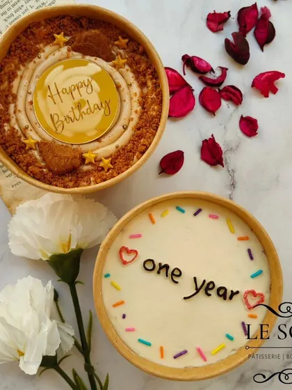 A duo of cake tubs for different celebrations. One is a Biscoff-flavored tub for a birthday, and the other is a simple "one year" anniversary cake with rainbow sprinkles.