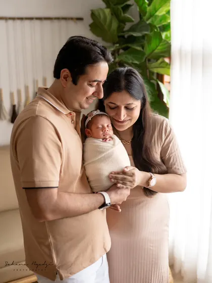 New parents looking down at their baby, held securely in their arms. The soft window light and their coordinated outfits create a harmonious and loving family portrait.