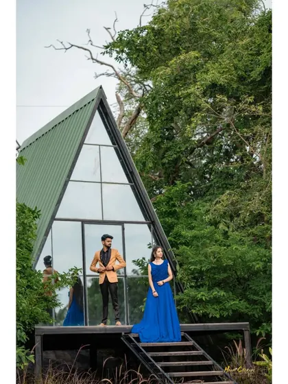 A wide shot showcasing a couple posing in front of a unique A-frame glass house, surrounded by nature. The bride's blue dress adds a touch of elegance to the modern, rustic setting.