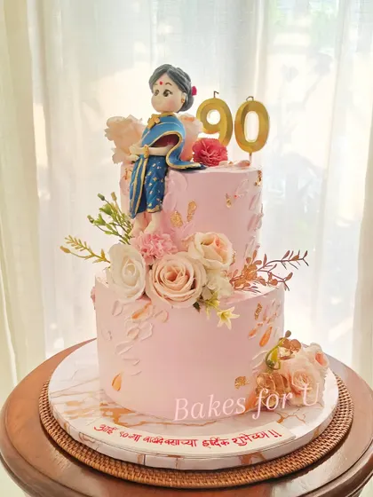 A truly special two-tier cake for a 90th birthday, featuring a handcrafted fondant figure of the matriarch in a beautiful blue saree. The cake is written in Marathi, adding a personal, local touch.