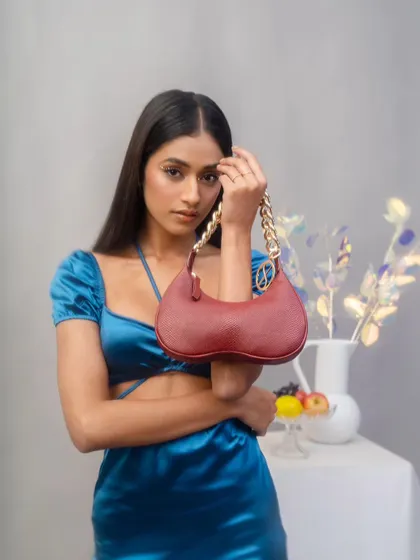 A model poses with a red Sarjaa handbag in a studio setting. The composition draws the eye directly to the product, highlighting its unique shape and gold chain detail.