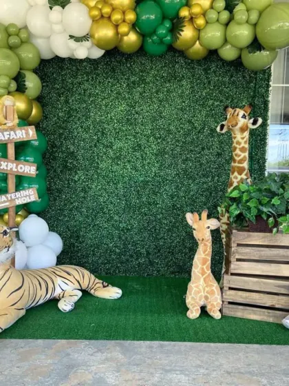 A safari-themed decoration featuring a lush green hedge wall as a backdrop. The scene is completed with realistic stuffed animals like a tiger and giraffes, and a green and gold balloon arch.