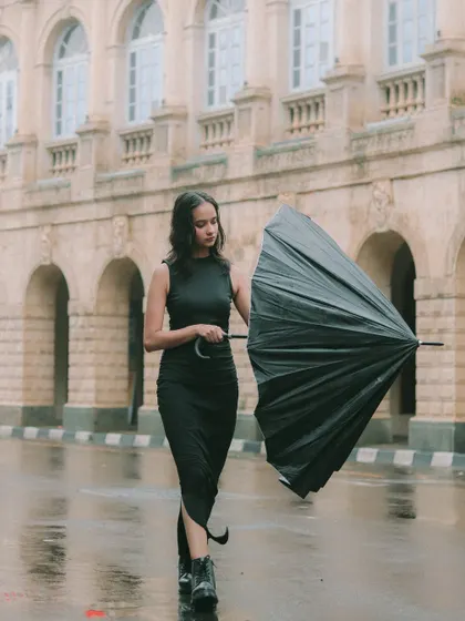 A woman walks through the rain in a black dress, umbrella in hand, against the backdrop of Mumbai's colonial architecture. A truly cinematic solo portrait.